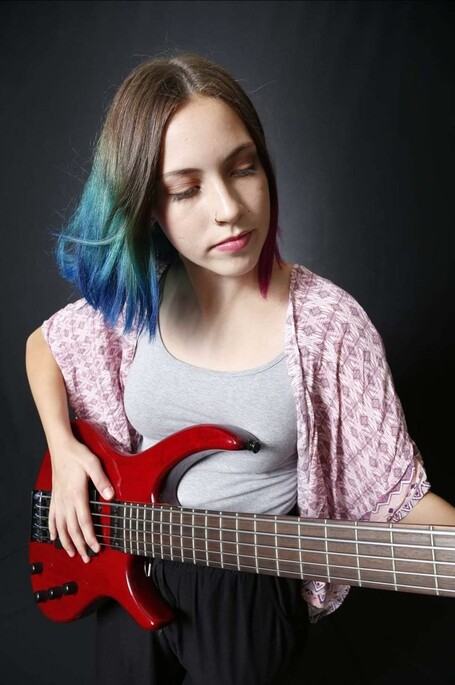 A girl with blue-tipped hair plays a guitar in front of a grey background.